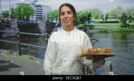 Die Chefin hält ein Tablett mit Muffins, Finger an die Lippen für Stille auf einer Straße neben einem Kanal; verspielte Geheimhaltung. Stockfoto