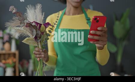 Floristin mit grüner Schürze hält Smartphone und Strauß aus getrockneten Gräsern und Eukalyptus im Blumengeschäft-Studio; Freude an kleinen Unternehmen. Stockfoto