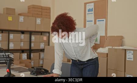 Frau hält sich im Gebäude auf den unteren Rücken, während sie sich auf den Schreibtisch neben gestapelten Kartonschachteln und Paketband stützt; kleine Unternehmen leiden. Stockfoto