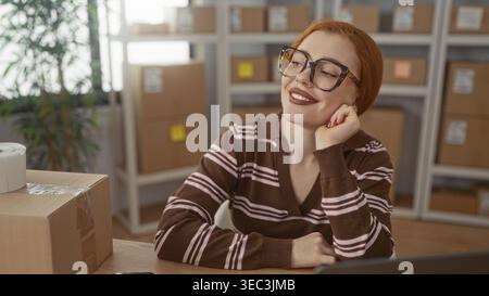 Rothaarige Frau mit Brille auf der Hand neben gestapelten Papppaketen in einem kleinen Verpackungsgebäude; unternehmerische Zufriedenheit. Stockfoto
