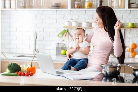 Mutter kocht gerne, während ihr Baby lächelt und in der Küche zu Hause spielt Stockfoto