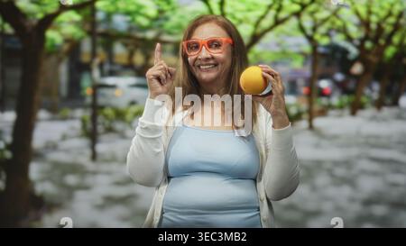 Eine Frau, die einen orangen Finger auf eine von Bäumen gesäumte Stadtstraße hält. Sie trägt eine orange Brille, ein hellblaues Tanktop und eine weiße Strickjacke Stockfoto