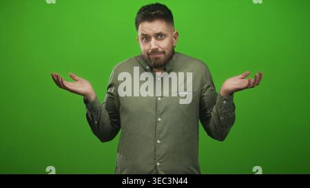 Der Mann zuckt mit den Schultern mit den Handflächen nach oben und geschlossenen Augen auf dem grünen Bildschirm des Studios, ein olivfarbenes Hemd sichtbar; Zweifel an Verwirrung. Stockfoto
