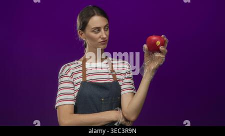 Frau in gestreiftem Hemd und Denim-Schürze mit rotem Apfel mit Handschuhen in lila Atelier; Hygieneinspektion. Stockfoto