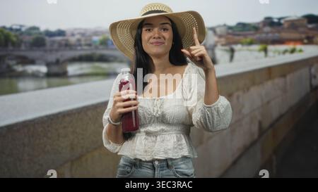 Junge Frau, die einen roten Saft in der Flasche hält und mit dem Zeigefinger auf einer Steinbrücke über einem Stadtfluss lächelt; glückliche Entspannung. Stockfoto