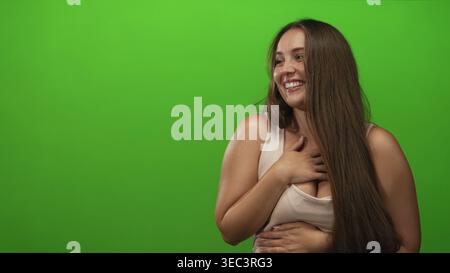 Lachende Frau mit Hand auf Brust und Hand auf Bauch mit Dekolleté und langen Haaren im grünen Atelier; Freude. Stockfoto