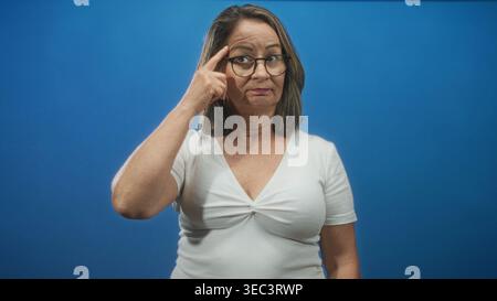 Hispanische Seniorin mit grauem Haar und Brille, die weiße Spitze trägt, Finger zum Tempel im Studio; nachdenkliche Reflexion. Stockfoto