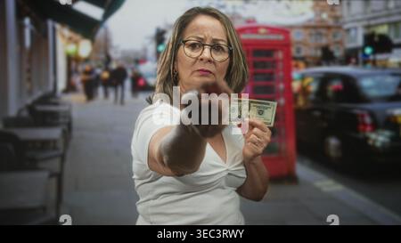 Hispanische Frau mit grauem Haar und Brille zeigt mit dem Finger, während sie auf der belebten Straße eine 20-Banknote hält; Ärger bei der Finanznachfrage. Stockfoto