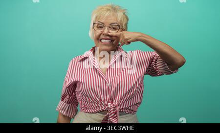 Eine ältere hispanische Frau mit Brille und einem gebundenen rot gestreiften Hemd zeigt den Zeigefinger auf ihre Nase, während sie vor einem türkisfarbenen Hintergrund in Studi lächelt Stockfoto