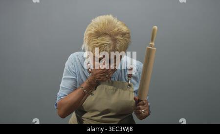 Frau kocht in Schürze, hält eine Nudelnadel und bedeckt ihren Mund mit der Hand, während sie im Studio klägt; Unwohlsein. Stockfoto