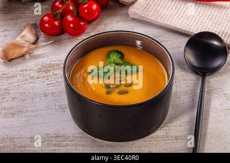 Kräftige, orange Kürbissuppe in dunkler Keramikschale. Garniert mit frischen Basilikumblättern und gerösteten Kürbiskernen. Warmes, einladendes Herbstessen, gesundes Essen Stockfoto