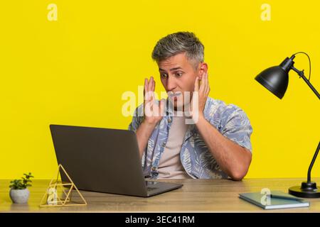 Ein Mann mittleren Alters liest unerwartete E-Mails auf einem Laptop am Tisch, zeigt einen Alarmfokus und lehnt sich dann plötzlich mit weit geöffneten Augen geschockt zurück. Junger Mann, der emotional auf Überraschung reagiert, auf gelbem Hintergrund. Stockfoto