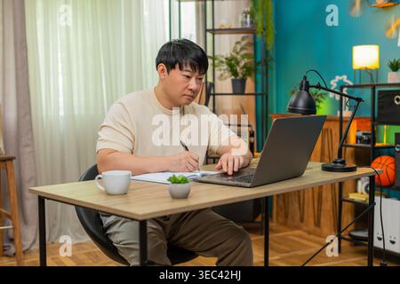 Asiatischer Mann, der zu Hause am Tisch sitzt, nimmt an einem Online-Kurs auf Laptop Teil, schreibt Notizen und erklärt der Kamera, wie sie sich zeigt. Der Chinese spricht selbstbewusst, mit ausdrucksstarken Gesten, um ansprechende Online-Lektion zu erstellen. Stockfoto