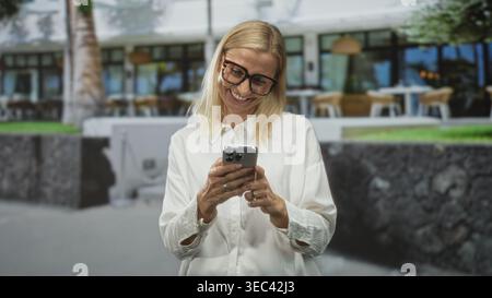 Frau mit Brille und weißem Hemd, die ihr Smartphone hält, den Bildschirm mit den Fingern klopft und lächelt, während sie vor dem Gebäude mit einem Café im Freien blickt Stockfoto