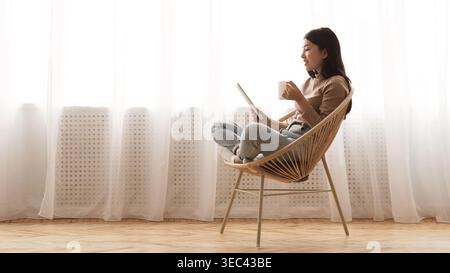 Frau, die sich in einem gemütlichen Stuhl mit einer Tasse Tee am Fenster in einem hellen Raum entspannt Stockfoto