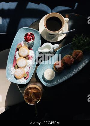 Blick von oben auf kleine süße Syrniki-Pfannkuchen mit frischem Joghurt und Heidelbeeren und Mini Syrniki-herzhafte Lachspancakes mit schwarzem Kaffee und einem Eis-Espresso-Tonic auf einem Tisch in einem sonnigen Café Stockfoto