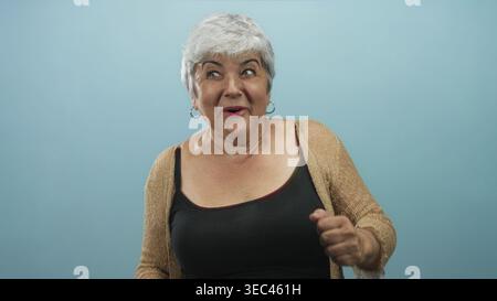 Frau, die Arme hebt und lächelt, mit freiliegenden Schultern und Creolen vor blauem Studiohintergrund, mit Strickjacke und Tanktop; Freude feiern Stockfoto