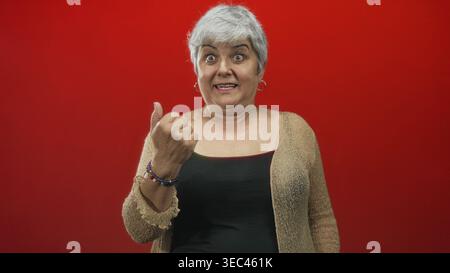 Frau zeigt mit gestreckter Hand und starkem Ausdruck mit dem Finger auf die Kamera, trägt Strickjacke und Tanktop vor rotem Studiohintergrund; beschimpft die Schuld. Stockfoto