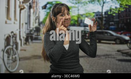 Frau zeigt Finger an Wange, während sie eine Kreditkarte auf einer kopfsteingepflasterten Straße hält; Vertrauen finanziert. Stockfoto
