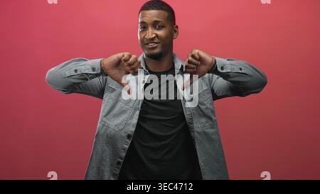 Der junge schwarze Mann trägt ein graues Denim-Hemd und ein schwarzes T-Shirt und zeigt die Daumen nach unten mit beiden Daumen im roten Studio-Hintergrund. Stockfoto