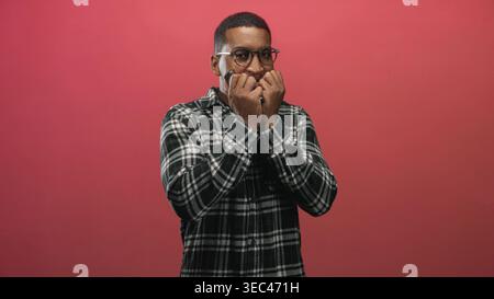 Mann mit Autoschlüsseln, Hand-zu-Mund-Geste, kariertes Hemd vor rotem Studio Hintergrund; Angst Zweifel Unsicherheit. Stockfoto