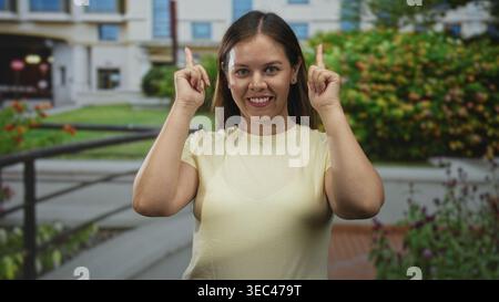 Die Frau zeigt beide Zeigefinger nach oben vor den Eingang des Gebäudes und lächelt in die Kamera; Optimismus wird ermutigt. Stockfoto