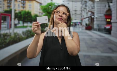 Frau mit weißer Kreditkarte zeigt Finger zum Kinn auf der geschäftigen Straße der Stadt; durchdachte Kaufentscheidung. Stockfoto