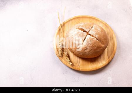 Runder, rustikaler hausgemachter Brotlaib mit einer staubigen Mehlplatte und Weizenohren auf einem Holzteller, auf einem hellgrauen Hintergrund, Representin Stockfoto
