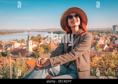 Frau mit Hut und Sonnenbrille genießt Panoramablick auf die Stadt und den Fluss im Herbst. Reisender weiblicher Tourist, der neue Ziele erforscht Stockfoto