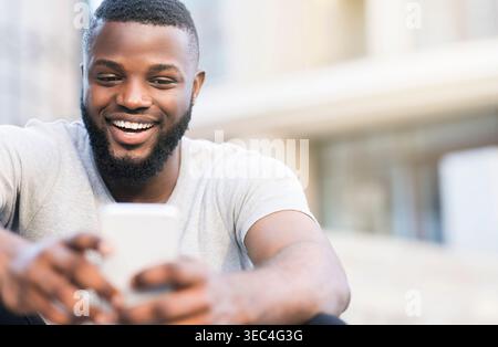 Junger Mann lächelt und benutzt das Smartphone, während er draußen in der städtischen Umgebung sitzt Stockfoto