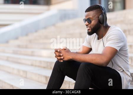 Junger Mann, der tagsüber Musik genießt, während er in einer Stadt auf Stufen sitzt Stockfoto