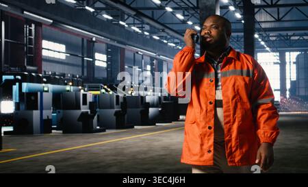 Der schwarze Bediener spricht über Walkie Talkie und überwacht Metallbearbeitungsgeräte in der Werkstatt, übernimmt die berufliche Verantwortung und technische Aufgaben im Zusammenhang mit dem Anlagenbetrieb. Stockfoto