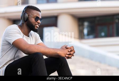 Mann, der auf Stufen sitzt, Kopfhörer und Sonnenbrille trägt und Musik im Freien in einer modernen Umgebung genießt Stockfoto