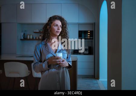 Junge Frau mit lockigen Haaren im Pyjama, die Kaffeetasse hält, die nachts in der modernen Küche steht. Die Dame isst Erfrischung, während sie wegblickt Stockfoto