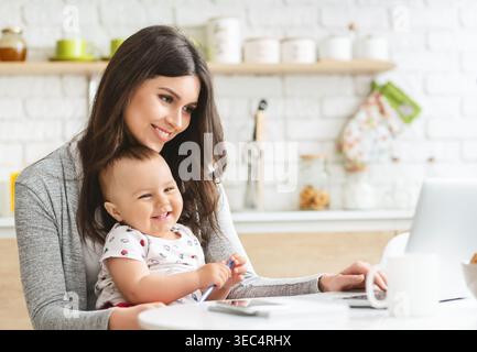Mutter genießt freudige Momente mit ihrem Baby, während sie am Küchentisch in heller Atmosphäre arbeitet Stockfoto