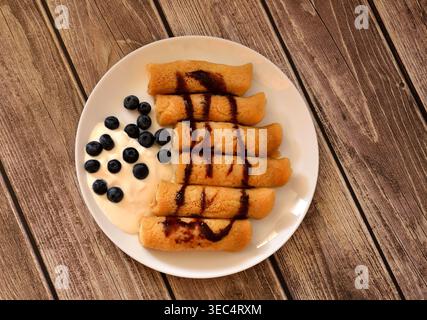 Ein Teller mit mehreren Pfannkuchen mit Sauerrahm und gefüllt mit Hüttenkäse, dekoriert mit Karamell und Heidelbeeren. Draufsicht, flach. Stockfoto