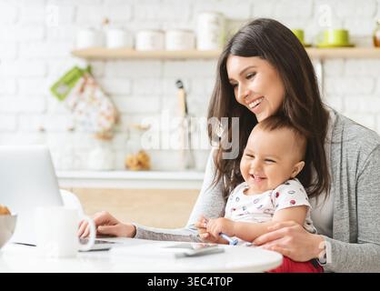 Mutter genießt die gute Zeit mit ihrem fröhlichen Baby, während sie zu Hause an einem Laptop arbeitet Stockfoto