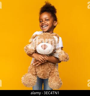 Glückliches Mädchen, das einen Teddybären vor einem hellen gelben Hintergrund hält, während er lächelt Stockfoto
