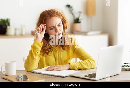 Junge Frau, die sich auf Aufgaben konzentriert, während sie mit Laptop und Notizblock an einem Tisch sitzt Stockfoto