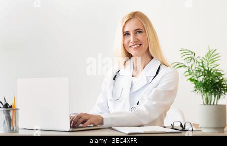 Junge Ärztin arbeitet an einem Schreibtisch mit einem Laptop und lächelt in einem modernen Büro Stockfoto