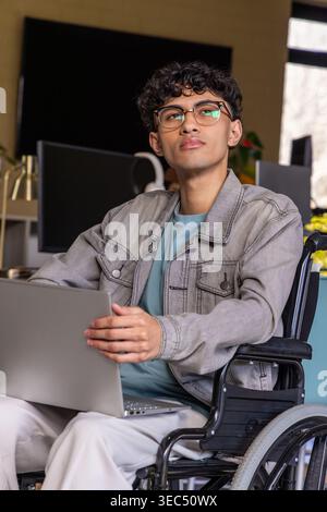 Asiatischer Mann, der im Rollstuhl am Schreibtisch sitzt und einen Laptop mit Jeansjacke und Brille hält Stockfoto