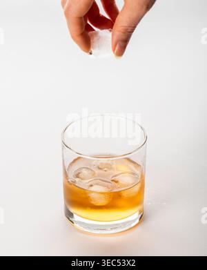 Nahaufnahme einer Frauenhand, die einen Eiswürfel in ein Whiskyglas wirft und Spritzer und Bewegung isoliert auf weißem Hintergrund erzeugt. Konzept des Luxusgetränks, Stockfoto