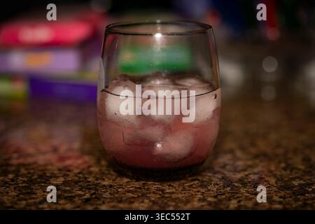 Nahaufnahme eines rosa Wodka-Cocktails mit Eiswürfeln in einem stiellosen Glas auf einer Granittheke mit einem unscharfen bunten Hintergrund bei schlechten Lichtverhältnissen Stockfoto