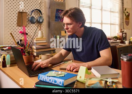 Junger Erwachsener männlicher kaukasischer Student, der auf einem Laptop studiert, Notizen am Schreibtisch macht, Kursarbeit leitet und Online-Lernen lernt. Ideal für Bildungsmarketing, E-Learning und Lifestyle-Kampagnen für Schüler Stockfoto