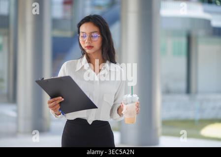 Geschäftsfrau, die ein Klemmbrett studiert und Eiskaffee in einem hellen Bürokorridor hält, hebt Konzentration, Planung und komponierte Professionalität hervor Stockfoto