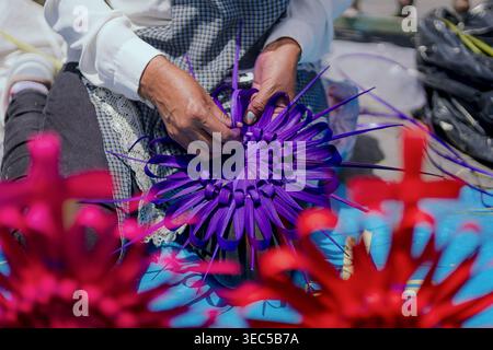 Nahaufnahme eines Handwerkers, der mit getrockneten und gefärbten Palmblättern eine komplizierte dekorative violette Blume webt, umgeben von anderen farbenfrohen Kreationen Stockfoto