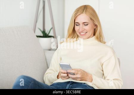 Frau lächelt, während sie ihr Smartphone zu Hause in einem modernen Wohnzimmer an einem sonnigen Tag benutzt Stockfoto