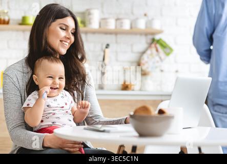 Mama genießt die gute Zeit mit ihrem glücklichen Baby, während sie zu Hause an einem Laptop arbeitet Stockfoto