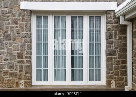 Großes, weiß gerahmtes Fenster mit vier Scheiben und Steinmauern, Hausaußenwand mit Dachrinne und Fallrohr, Wohnarchitektur und Gebäude Stockfoto