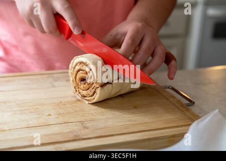 Nahaufnahme der Hände Schneiden von Zimtbrötchen-Teig auf Holzbrett in der Hausküche, Backprozess und hausgemachtes Lebensmittelzubereitungskonzept Stockfoto